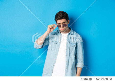 Confident macho man put on sunglasses, looking cool and sassy, standing over blue background Confident macho man put on sunglasses, looking cool and sassy, standing over blue background 82347018