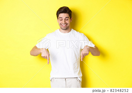 Young handsome man pointing and looking down, checking out promo offer, standing over yellow background 82348252