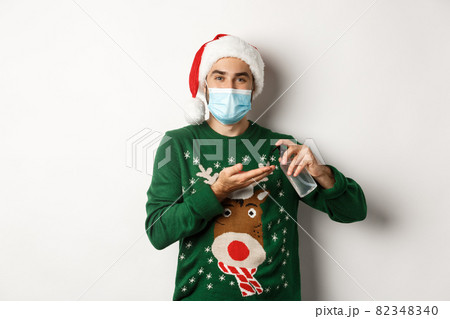 Concept of covid-19 and Christmas holidays. Caucasian man in face mask and sweater using antiseptic, clean hands with sanitizer, standing over white background Concept of covid-19 and Christmas holidays. Caucasian man in face mask and sweater using antiseptic, clean hands with sanitizer, standing over white background 82348340