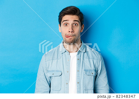 Close-up of young man making funny expressions, showing tongue and looking at camera, standing over blue background Close-up of young man making funny expressions, showing tongue and looking at camera, standing over blue background 82348347