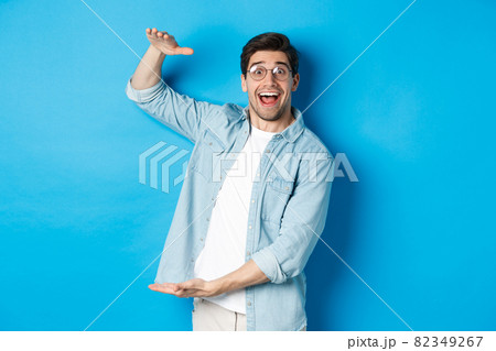 Happy man in glasses showing big size object, shaping large box, standing over blue background and smiling 82349267