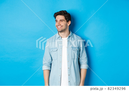 Handsome smiling adult man in casual outfit, smiling and looking left at promo offer, standing against blue background 82349296
