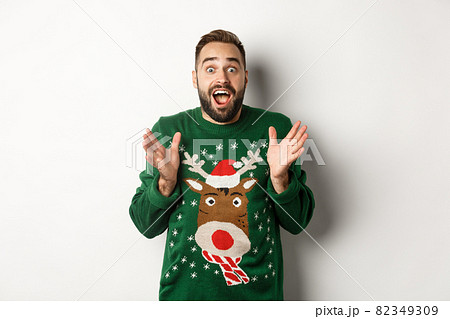 New year celebration and winter holidays concept. Happy and surprised bearded guy looking amazed, catching something, standing in funny Christmas sweater, white background New year celebration and winter holidays concept. Happy and surprised bearded guy looking amazed, catching something, standing in funny Christmas sweater, white background 82349309