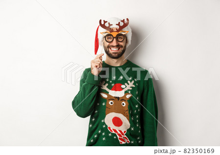 Christmas and holidays concept. Smiling beared man in Santa hat looking happy, holding party mask for New Year, celebrating xmas, standing over white background Christmas and holidays concept. Smiling beared man in Santa hat looking happy, holding party mask for New Year, celebrating xmas, standing over white background 82350169