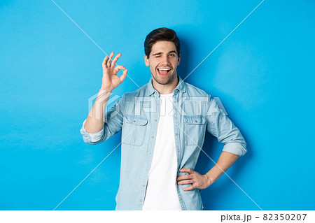 Confident adult man winking and showing okay sign, approve something good, standing against blue background Confident adult man winking and showing okay sign, approve something good, standing against blue background 82350207