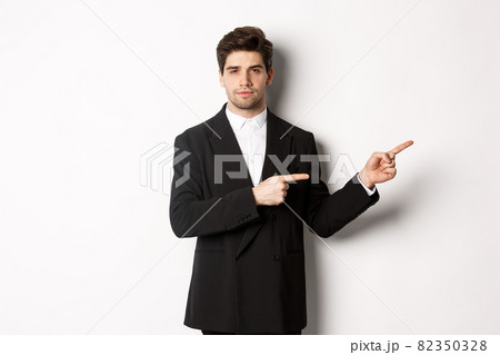 Image of handsome businessman in black suit, pointing fingers right and looking at camera, standing against white background Image of handsome businessman in black suit, pointing fingers right and looking at camera, standing against white background 82350328