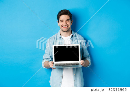 Handsome young man introduce product on laptop screen, showing computer and smiling, standing over blue background 82351966