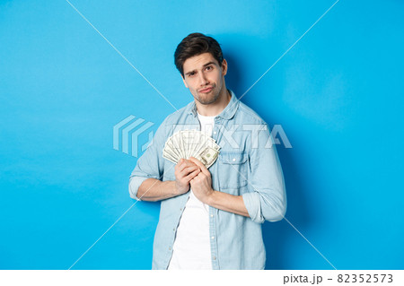 Handsome young man keeping money to himself, smiling and looking greedy, standing over blue background 82352573