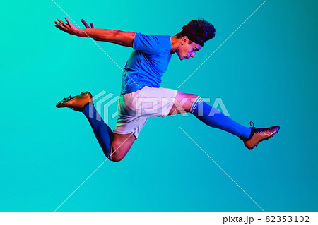 Full-length horizontal portrait of young man, soccer football player training isolated on blue background. In a jump Full-length horizontal portrait of young man, soccer football player training isolated on blue background. In a jump 82353102
