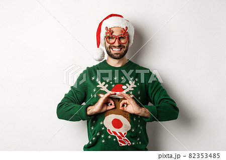 Christmas holidays, celebration concept. Happy guy in Santa hat and party glasses making fun of funny sweater, standing over white background 82353485