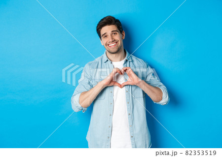 Handsome man smiling, showing heart gesture and looking at camera, saying I love you, standing against blue background 82353519