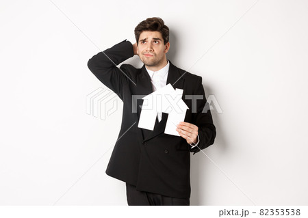 Image of indecisive businessman thinking, holding house maket and looking at upper left corner doubtful, standing against white background 82353538
