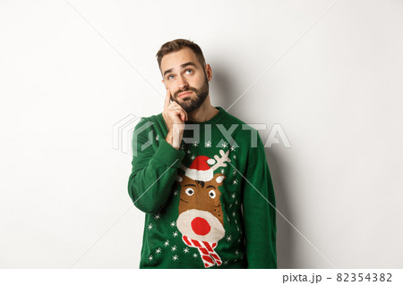 New Year, holidays and celebration. Sad young man with beard, wearing green sweater, showing a teardrop on his face and looking up upset, standing over white background 82354382