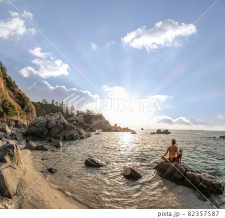 Girl with dog sitting on a rock next to the beach and meditating against sunset over the sea. Calabria paradise in Italy 82357587