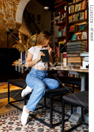 Pretty smiling joyfully female with fair hair, dressed casually, looking with satisfaction at camera, being happy. Full body shot of good-looking beautiful woman sitting against bookshelf wall.  82359771