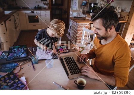 Mid adult father with small son working indoors at home, painting pictures and home office concept. Mid adult father with small son working indoors at home, painting pictures and home office concept. 82359890