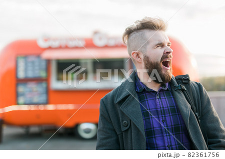 Bearded man in the street yawns with opened mouth stands at a food truck background. Sleepy people and lazy concept. Bearded man in the street yawns with opened mouth stands at a food truck background. Sleepy people and lazy concept. 82361756