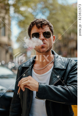 portrait of a sexy brutal man, in sun glasses. he exhales smoke and smokes a cigarette in public place of a city portrait of a sexy brutal man, in sun glasses. he exhales smoke and smokes a cigarette in public place of a city 82361973