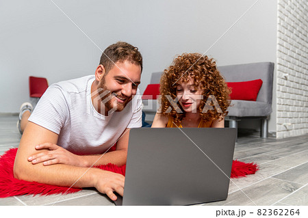 A Young couple lovers or friends laying on a red pluffy carpet and using laptop. 82362264
