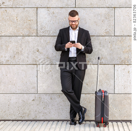 Confident businessman in formal outfit with suitcase using mobile phone outdoors, waiting for taxi 82363706