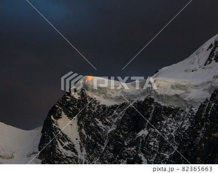 Darkness mountain landscape with great snowy mountain lit by dawn sun among dark clouds. Awesome alpine scenery with high mountain pinnacle at sunset or at sunrise. Big glacier on top in orange light. Darkness mountain landscape with great snowy mountain lit by dawn sun among dark clouds. Awesome alpine scenery with high mountain pinnacle at sunset or at sunrise. Big glacier on top in orange light. 82365663