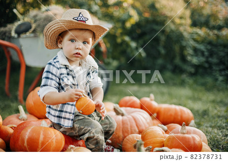A little cowboy boy in a hat and a plaid shirt is sitting on a pile of pumpkins in the garden and holding a small pumpkin in his hands. Autumn, the time of harvest 82378731