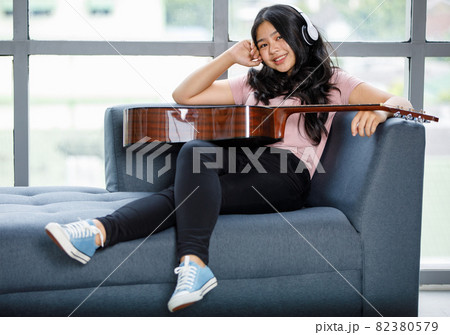 Portrait shot of young teenager enjoying playing the acoustic guitar and wearing a headset. Junior guitarist sitting on the couch and holding an instrument while posing and looking at the camera Portrait shot of young teenager enjoying playing the acoustic guitar and wearing a headset. Junior guitarist sitting on the couch and holding an instrument while posing and looking at the camera 82380579