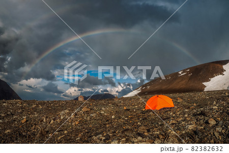 Panorama with a rainbow and an orange tent in the mountains. Atmospheric alpine landscape with snowy mountains with rainbow in rainy and sunny weather. 82382632