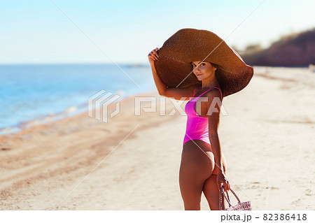 Back view of girl in sunhat and bikini on her perfect body walks sea white sand while holds handbag  82386418