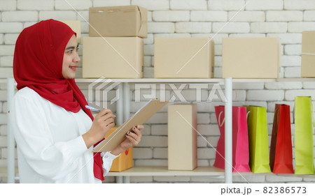 Portrait shot of cute smiling young teenage Muslim woman wearing red hijab standing while holding an order checklist to checking parcels carefully before delivery with postage boxes background 82386573