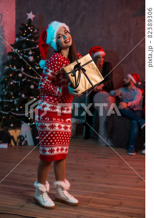 Portrait of a young woman with a gold giftbox at home on the foreground. Beautiful brunette in a Santa hat, red costume with deers Portrait of a young woman with a gold giftbox at home on the foreground. Beautiful brunette in a Santa hat, red costume with deers 82389306