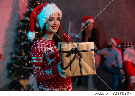 Portrait of a young woman with a gold giftbox at home on the foreground. Beautiful brunette in a Santa hat, red costume with deers Portrait of a young woman with a gold giftbox at home on the foreground. Beautiful brunette in a Santa hat, red costume with deers 82389307