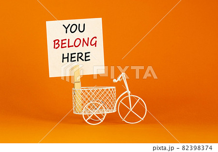 You belong here symbol. Wooden clothespin with white paper. Words you belong here. Miniature white bicycle. Beautiful orange background. Business and you belong here support concept, copy space. You belong here symbol. Wooden clothespin with white paper. Words you belong here. Miniature white bicycle. Beautiful orange background. Business and you belong here support concept, copy space. 82398374