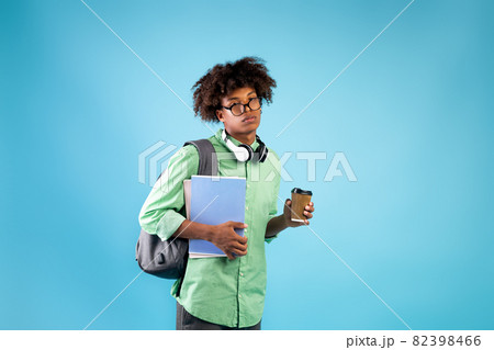 Exhausted male student with notebooks, takeaway coffee and backpack looking at camera on blue studio background 82398466