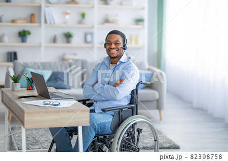 Positive paraplegic black man in wheelchair wearing headset, posing in front of laptop computer at home Positive paraplegic black man in wheelchair wearing headset, posing in front of laptop computer at home 82398758