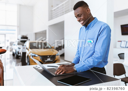 Positive black car salesman using laptop computer at work desk in auto dealership, copy space 82398760