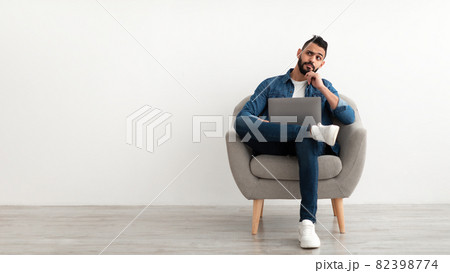 Thoughtful young Arab guy using laptop, sitting in armchair, looking aside at empty space against white studio wall 82398774