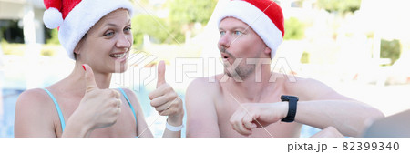 Young woman and man in santa claus hats in pool invites thumbs up gesture and smiles into laptop Young woman and man in santa claus hats in pool invites thumbs up gesture and smiles into laptop 82399340