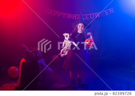 Funny child girl in witch costume for Halloween with pumpkin Jack and dog. 82399726