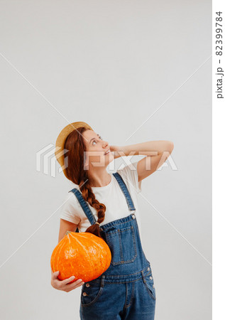 Farmer woman in overalls holds large pumpkin and looks up holding a wicker hat. Farmer woman in overalls holds large pumpkin and looks up holding a wicker hat. 82399784