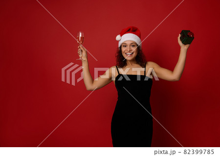 Elegant Hispanic woman wearing Santa hat holding champagne flute with sparkling wine and a gift box smiles toothy smile posing over red background with copy space. Merry Christmas, Happy New Year Elegant Hispanic woman wearing Santa hat holding champagne flute with sparkling wine and a gift box smiles toothy smile posing over red background with copy space. Merry Christmas, Happy New Year 82399785