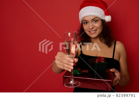 Cheerful African American woman with beautiful toothy smile, holds Christmas gift boxes wrapped in glitter red green gift paper and shows champagne flute with sparkling wine to camera. Copy space 82399791
