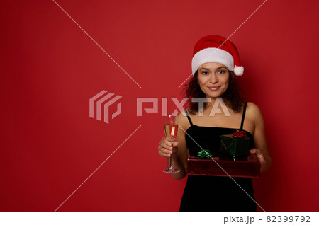 Beautiful Hispanic woman holds Christmas gift boxes wrapped in glitter red green gift paper and champagne flute with sparkling wine, poses on red background, cute smiling looking at camera. Copy space 82399792