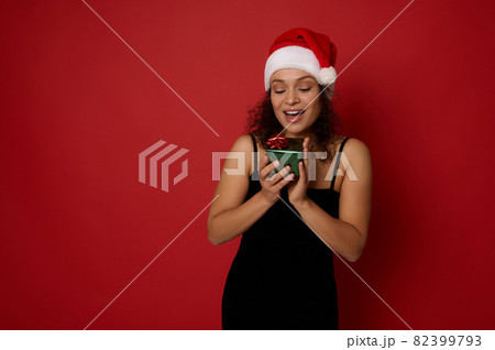 Happy Hispanic beautiful woman feeling happiness looking at small beautiful Christmas gift box in her hands, isolated over red background with copy space for ad. New Year and Merry Christmas concept. Happy Hispanic beautiful woman feeling happiness looking at small beautiful Christmas gift box in her hands, isolated over red background with copy space for ad. New Year and Merry Christmas concept. 82399793