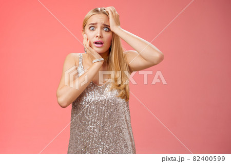 Shocked frightened insecure panicking woman blond hairstyle in silver dress touch head pop eyes afraid scared seing terrifying crime standing stupor speechless gasping shook, red background 82400599