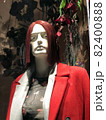 Woman's mannequin on the window of designer clothing store in Cyprus. Mannequin is wearing red jacket and translucent top. On the head is a burgundy wig with a bob hairstyle. Plants in the background 82400888