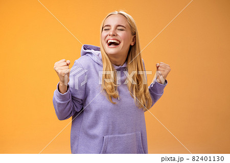 Cute happy triumphing blond european girl in hoodie yelling yes accomplish dream lifetime clench fists happily dancing celebrating victory success achieve goal, standing orange background 82401130