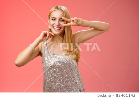 Charismatic funny amused charming blond girl 25s having fun dancing red background in silver trendy glittering party dress show peace disco gesture near eye smiling laughing carefree 82401142
