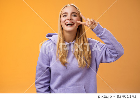 Attractive friendly carefree millennial blond girl in purple hoodie having fun friends laughing joyfully show peace victory hippie sign adore perfect chilly spring weather, orange background 82401143