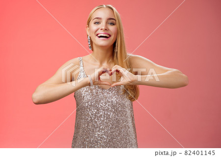 Adorable happy charming lovely blond european girl in silver dress express love positivity show heart gesture confess sympathy passionate feelings towards boyfriend smiling broadly, red background 82401145